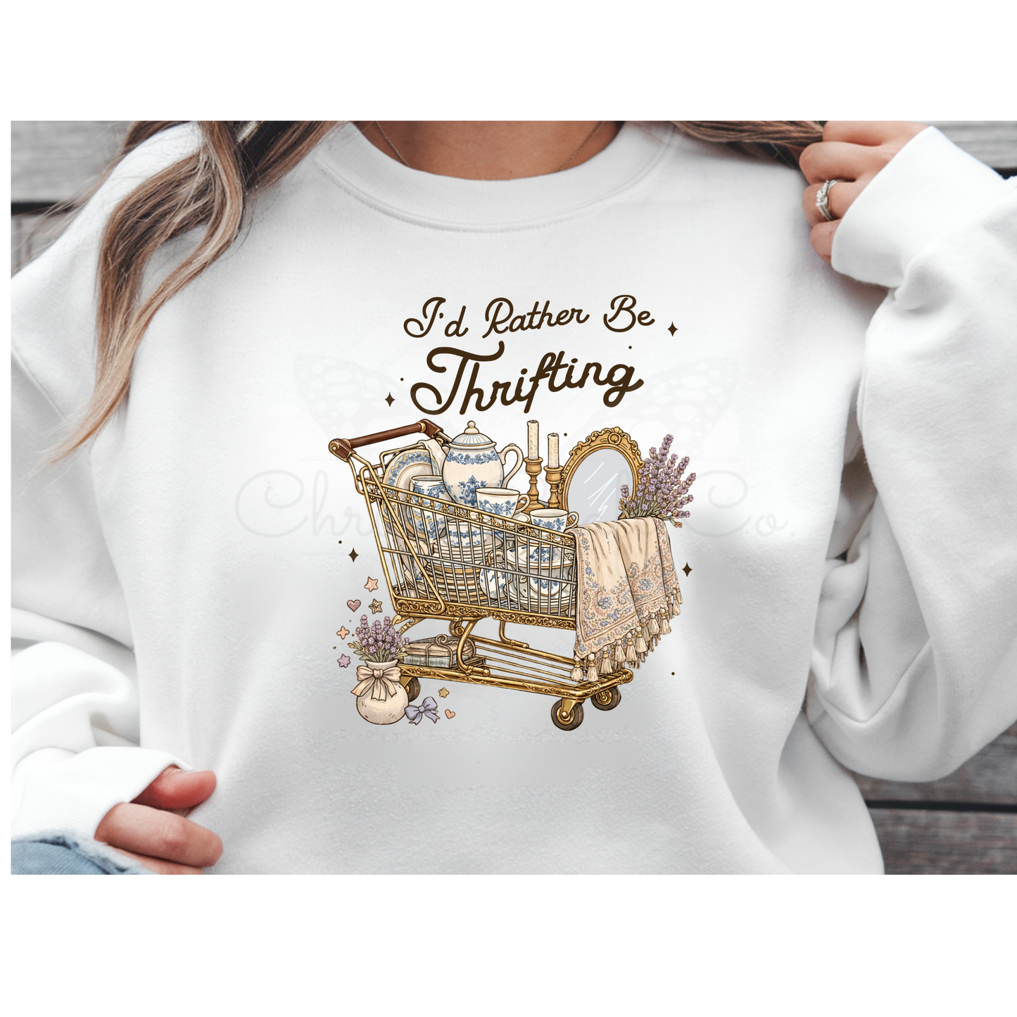 Mockup of a white sweatshirt featuring a vintage-style "I'd Rather Be Thrifting" DTF transfer. The design shows a distressed, ornate shopping cart filled with a teapot, books, and lavender flowers in a retro shabby chic aesthetic.