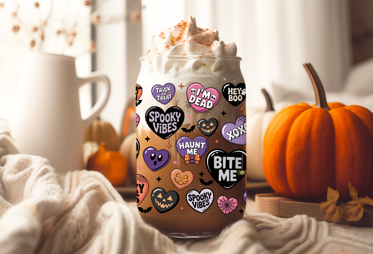 MOCKUP OF Clear glass 16oz can tumbler with a bamboo lid and glass straw, decorated with a pastel Halloween conversation hearts UVDTF wrap featuring phrases like Spooky Babe and Bite Me, displayed alongside iced drinks and fall decorations.