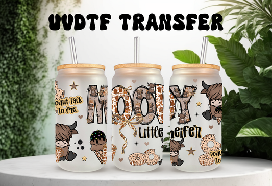 A UVDTF glass can wrap mockup featuring three 16oz frosted glass cans with bamboo lids and straws. The design displays a highland cow theme with the text "Moody Little Heifer" in cow-print lettering and "Donut Talk To Me." The graphic includes illustrations of highland cows, chocolate-dipped donuts with sprinkles, and small gold stars against a white background with green leafy plants.