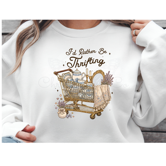 Mockup of a white sweatshirt featuring a vintage-style "I'd Rather Be Thrifting" DTF transfer. The design shows a distressed, ornate shopping cart filled with a teapot, books, and lavender flowers in a retro shabby chic aesthetic.