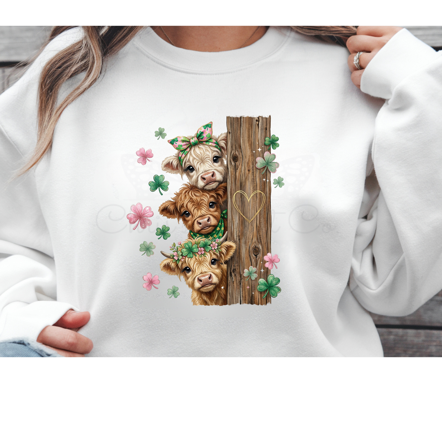 Three stacked Highland Cows wearing shamrock headbands next to a rustic wooden fence with pink flowers, vintage St. Patrick's Day DTF transfer on a white sweatshirt.
