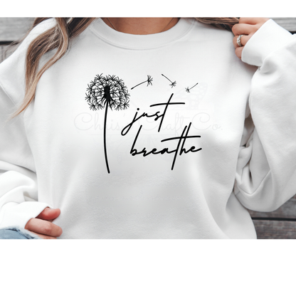 Minimalist black "Just Breathe" script next to a delicate dandelion flower with seeds blowing in the wind, shown as a DTF transfer on a white sweatshirt.