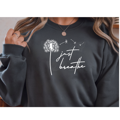 Minimalist WHITE "Just Breathe" script next to a delicate dandelion flower with seeds blowing in the wind, shown as a DTF transfer on a white sweatshirt.