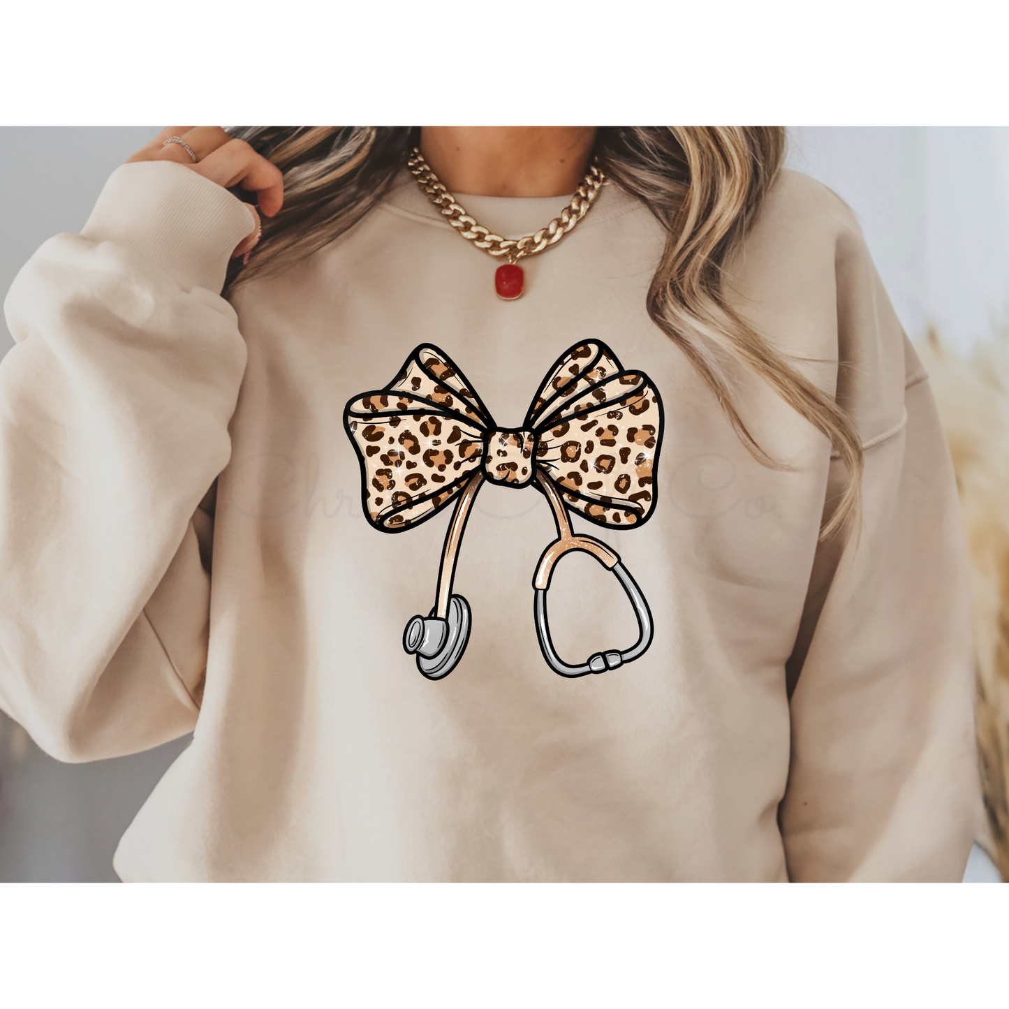 Mockup of a tan/beige sweatshirt featuring a leopard print bow tied to a stethoscope. The design is a high-quality DTF transfer centered on the chest.
