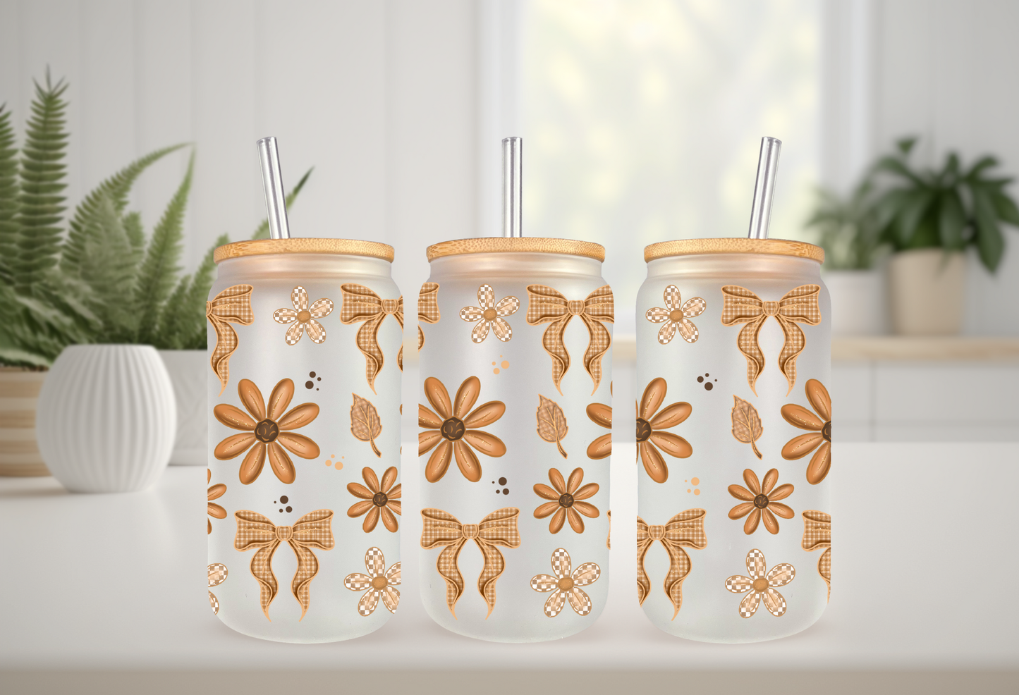 A mockup showcasing three frosted 16oz glass cans with bamboo lids and straws, decorated with orange daisy flowers and tan checkered bow UV DTF transfers