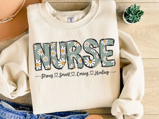 White sweatshirt featuring "NURSE" in daisy floral print with "Strong, Smart, Caring, Healing" text on a wooden background.